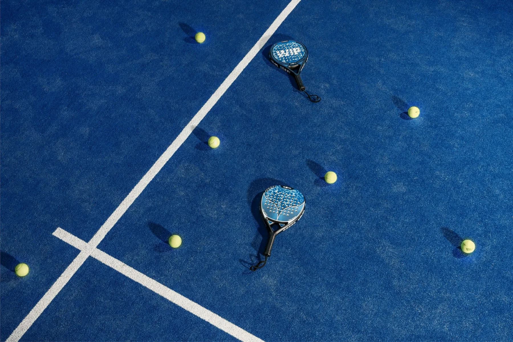 A padel court from directly above: blue surface, white lines, rackets and balls