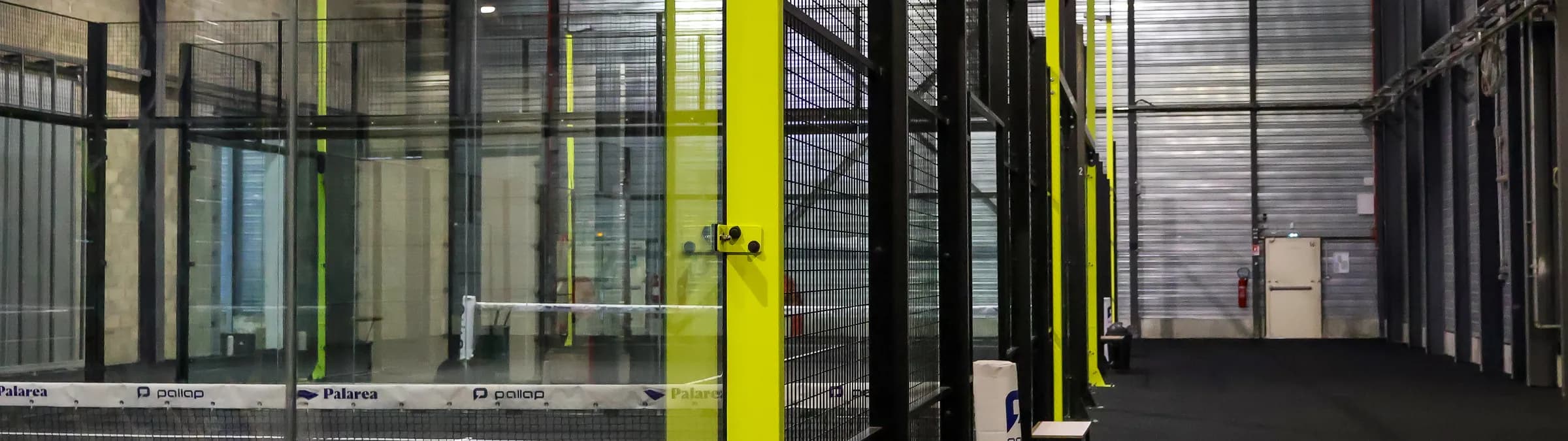An indoor padel hall with black frames, glass walls and yellow structural posts