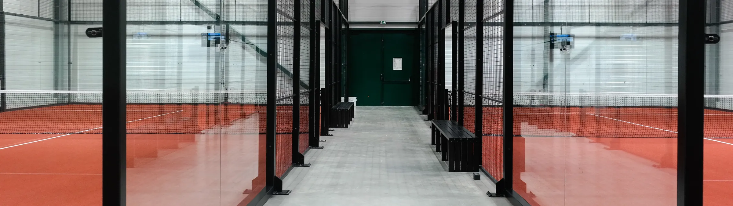An indoor padel hall, glass courts with terracotta turf either side of a central aisle