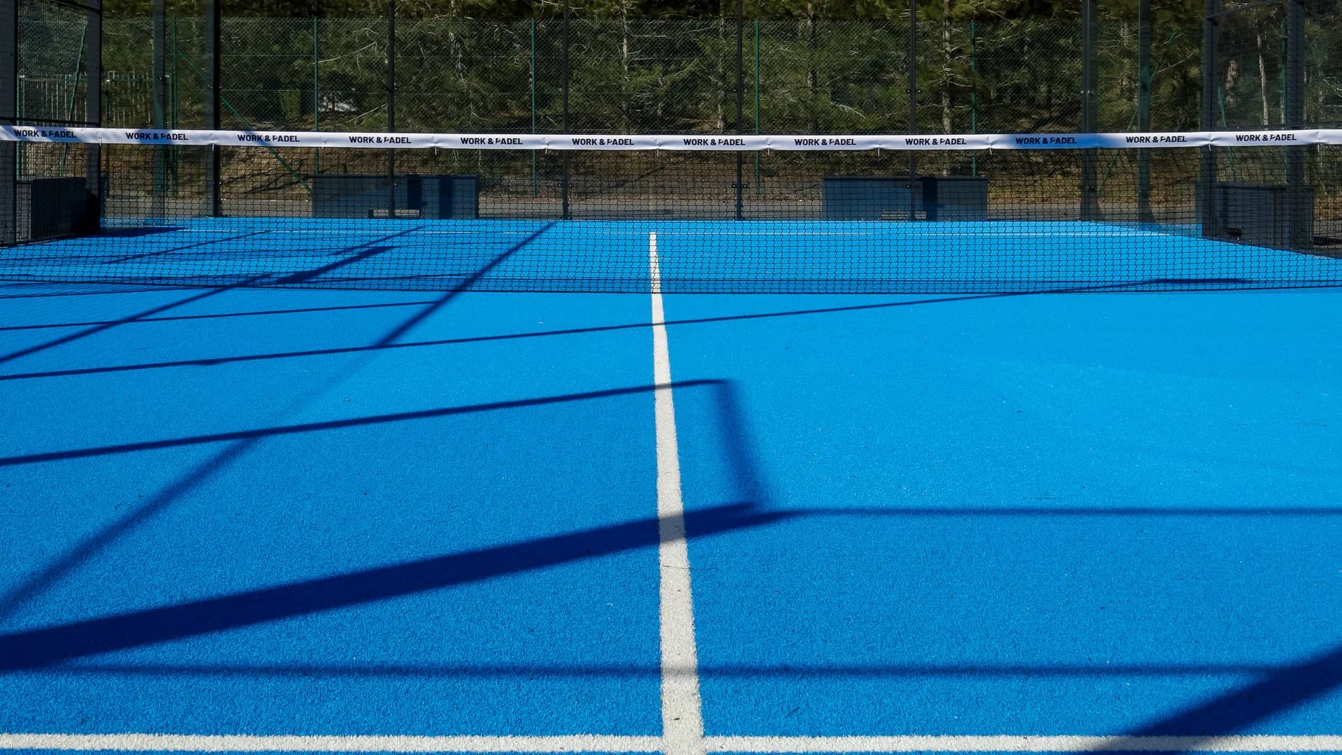 The net and white service line of a blue padel court in sunlight