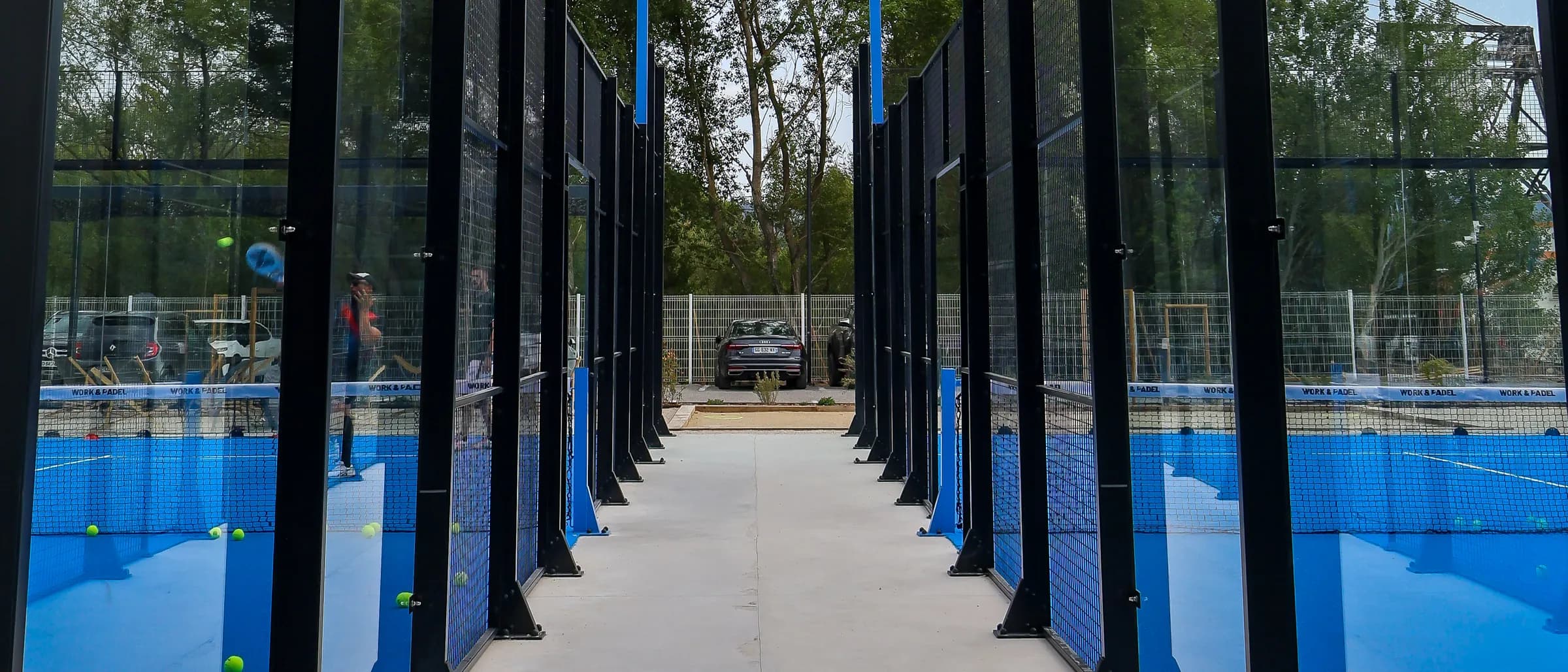 A walkway between two rows of blue padel courts with glass walls