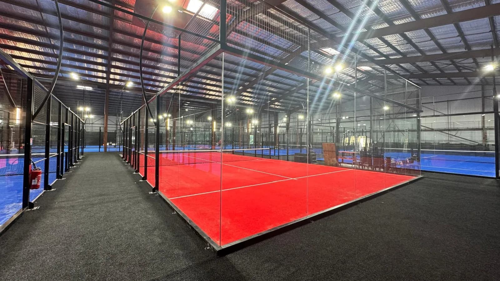 A full panoramic padel court with red turf and frameless glass walls in an indoor hall