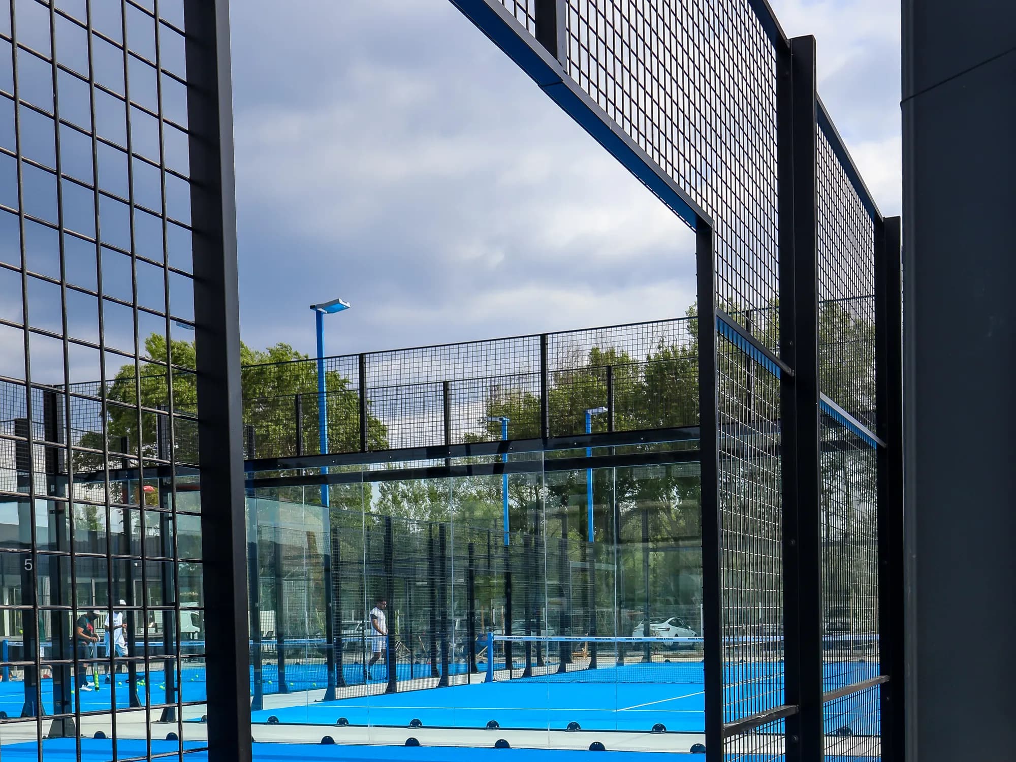 Players on a blue padel court with glass walls, seen through the black frame of the next court