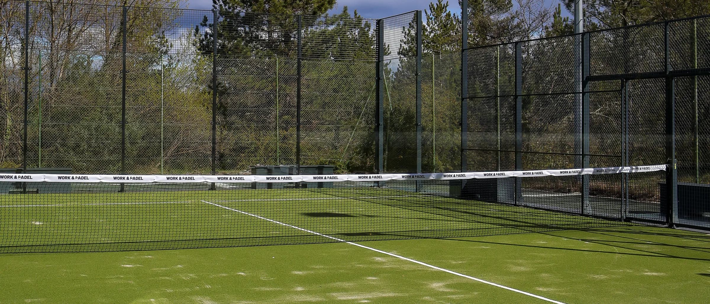 A green padel court with its net and floodlights, trees behind