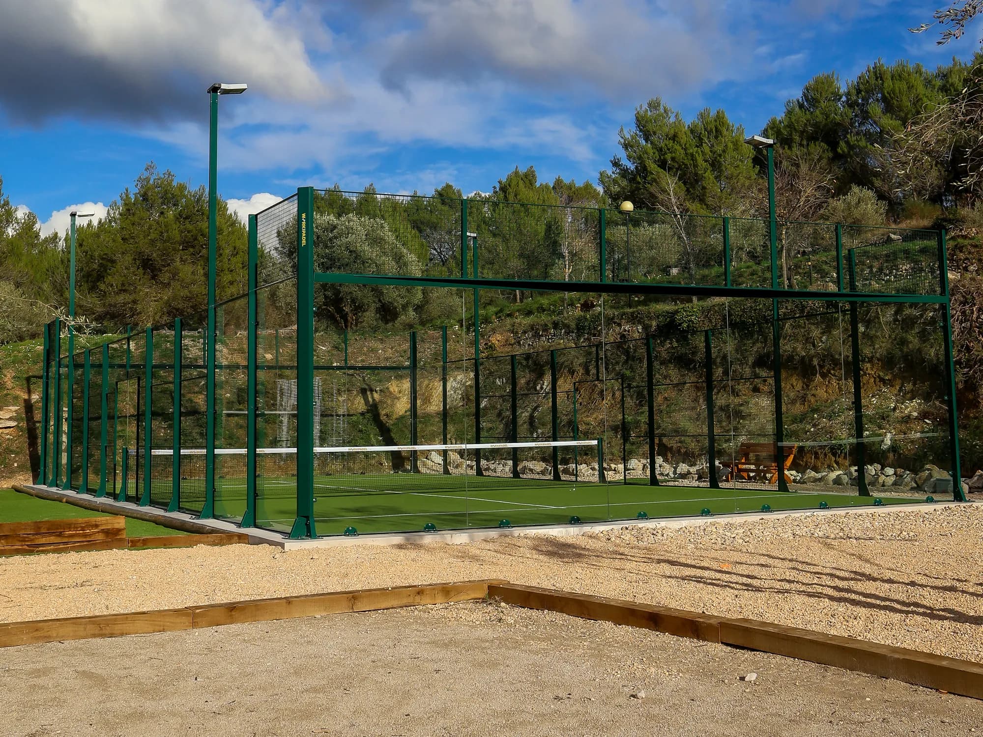 An outdoor padel court with green turf and a green steel frame among pine trees