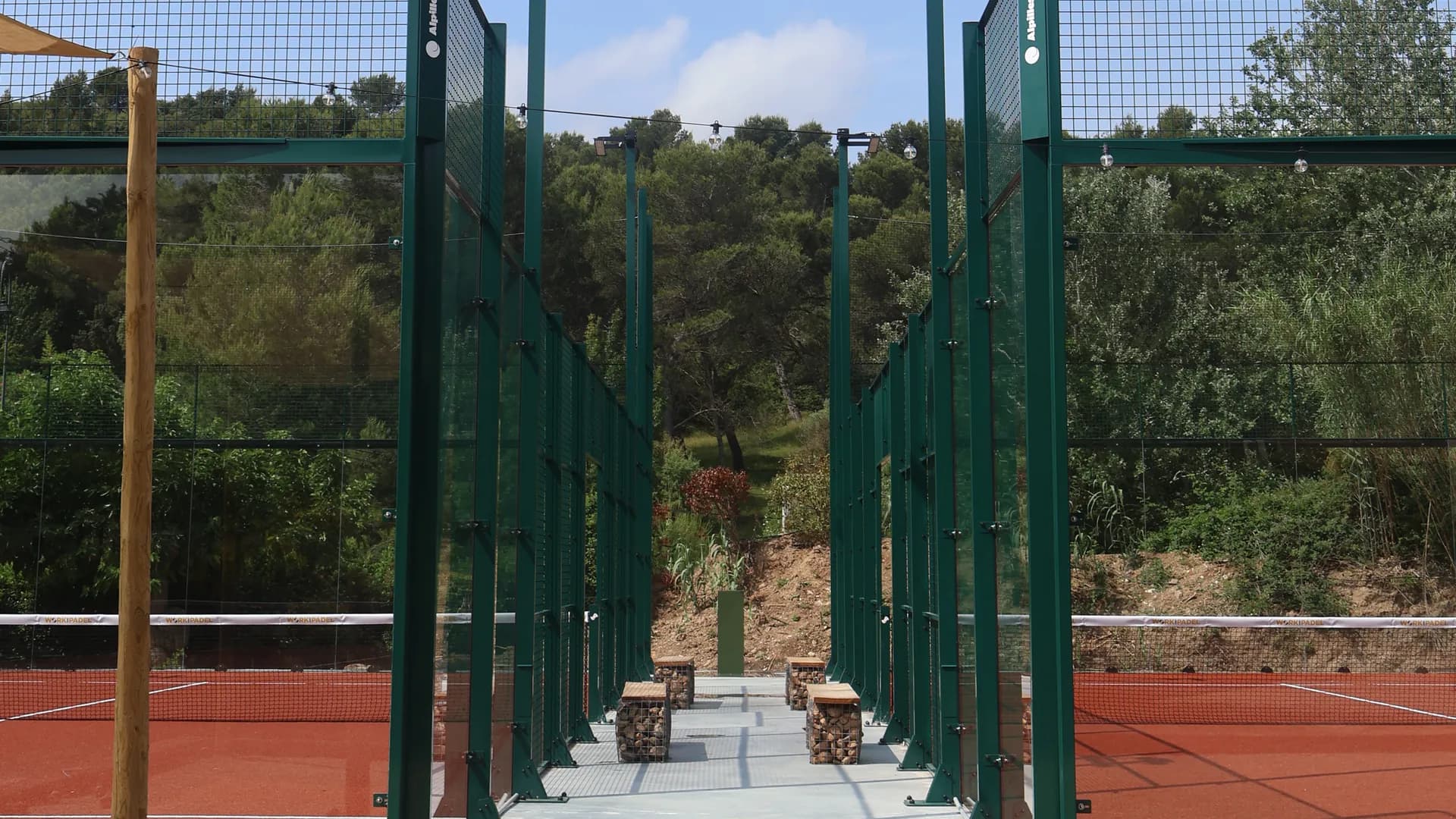 Two panoramic padel courts with green frames and terracotta turf, a walkway between them